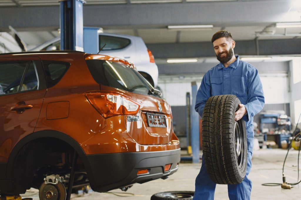 Lächelnder Mechaniker im blauen Overall hält einen Reifen vor einem orangen SUV
