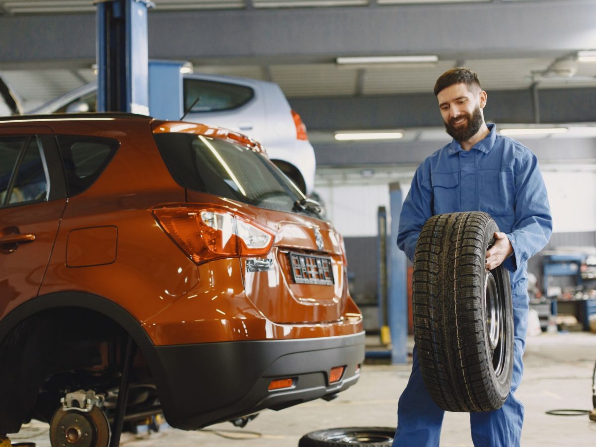 Lächelnder Mechaniker im blauen Overall hält einen Reifen vor einem orangen SUV